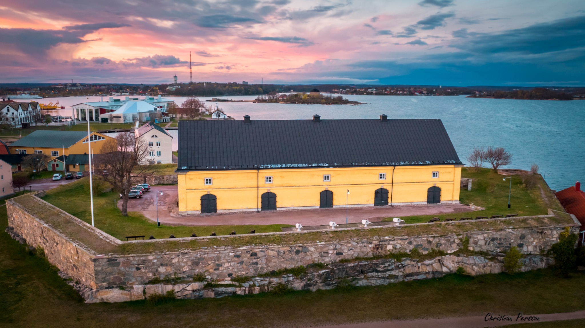 Flygbild av en gul byggnad bakom en stenmur vid havet i kvällsljus
