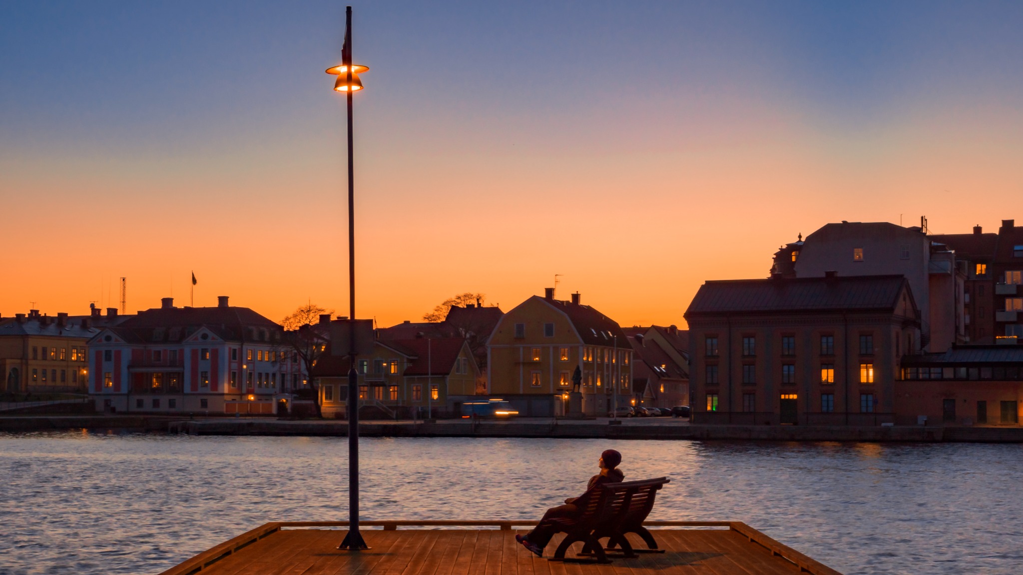 En person på en bänk vid vattnet med stadens hus mot en orange kvällshimmel